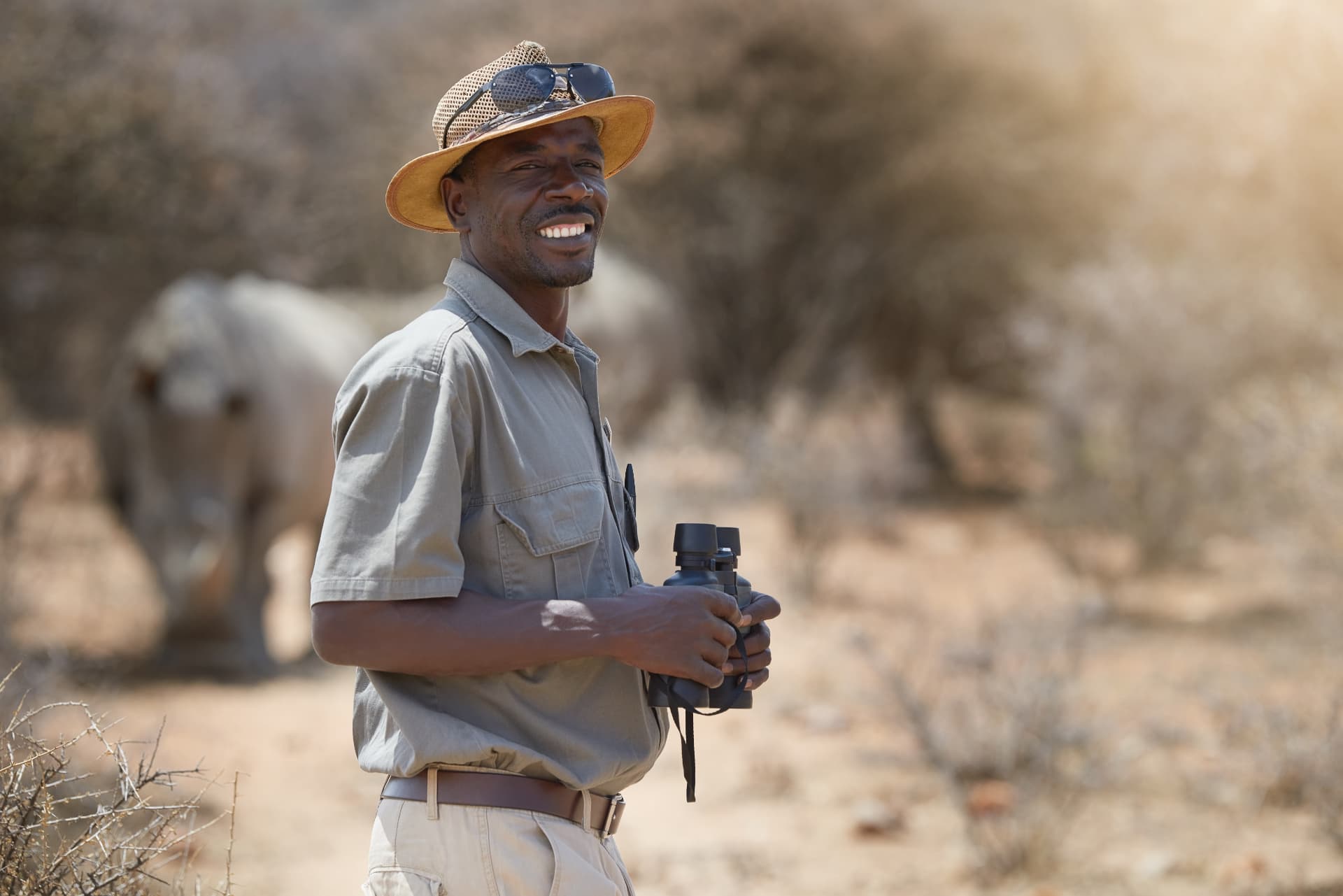 Smiling East African safari guide with binoculars, rhino grazing behind him